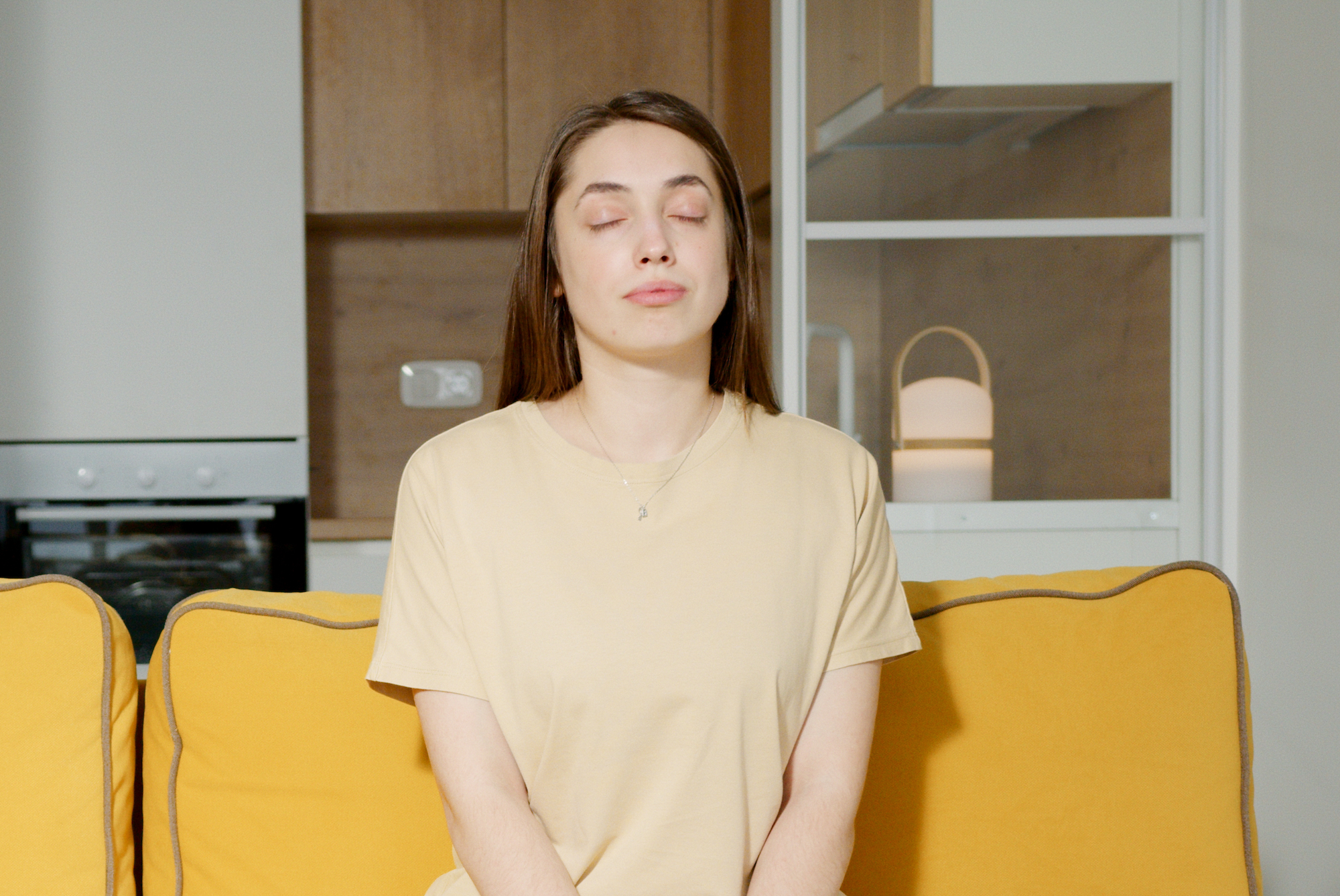 A person sits on a yellow couch in their home with the kitchen in the background. Their eyes are closed with their hands in their lap, as if meditating.
