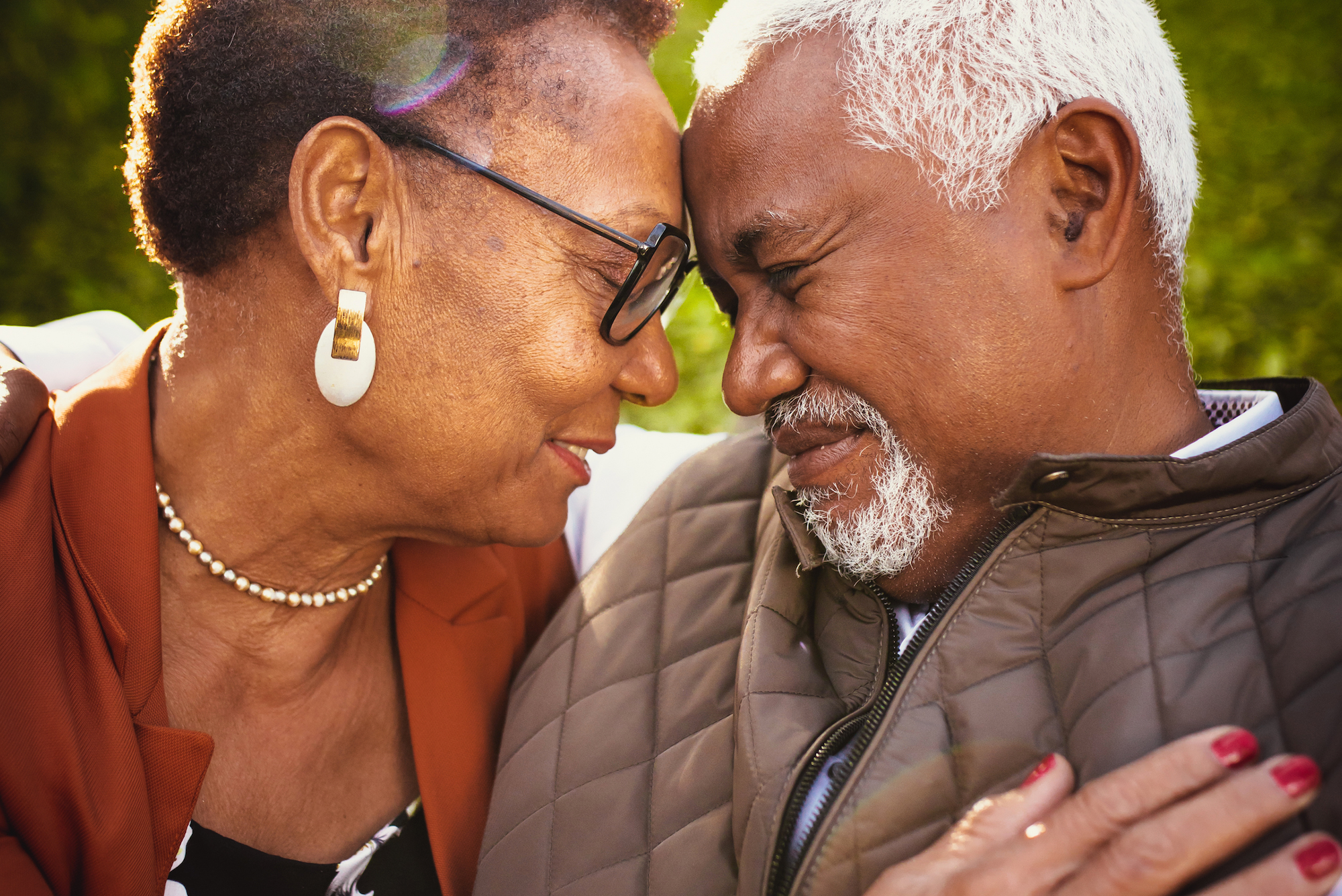 An elderly African American couple smiles and sits closely with their foreheads touching.