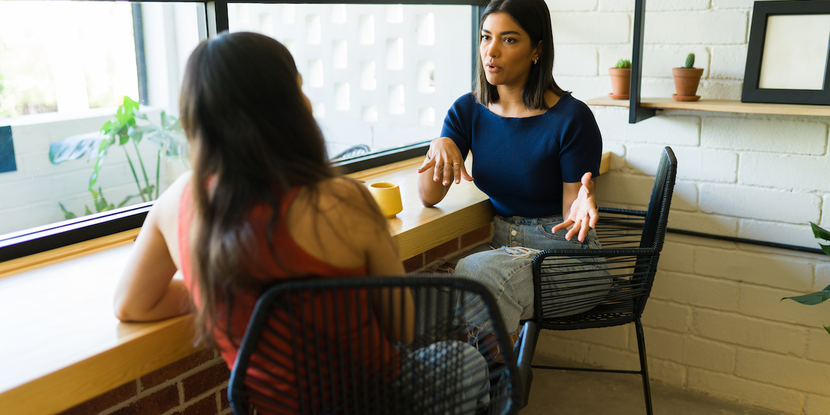 Two people sit at a counter with coffee mugs and hold a conversation