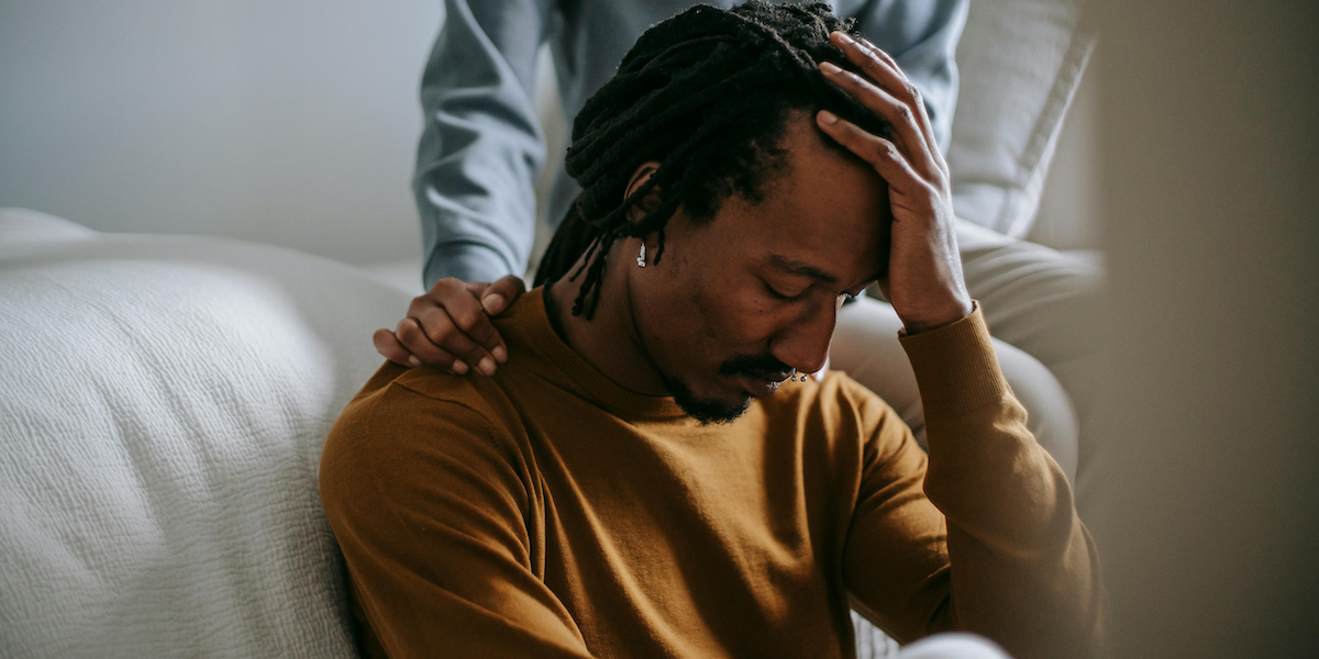 A Black adult with dreads, earrings, and a septum piercing sits in on a couch in sorrow with his head in his hands, while another person rests their hand on their shoulder.