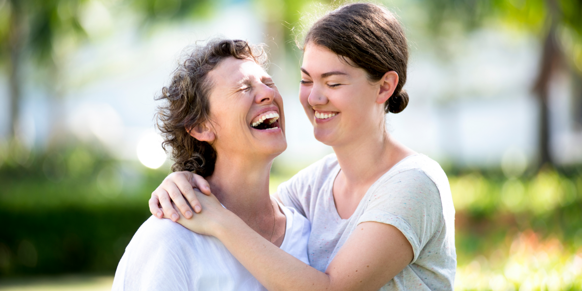 A mother and teenage daughter wear T-shirts and laugh together under the shade of trees.