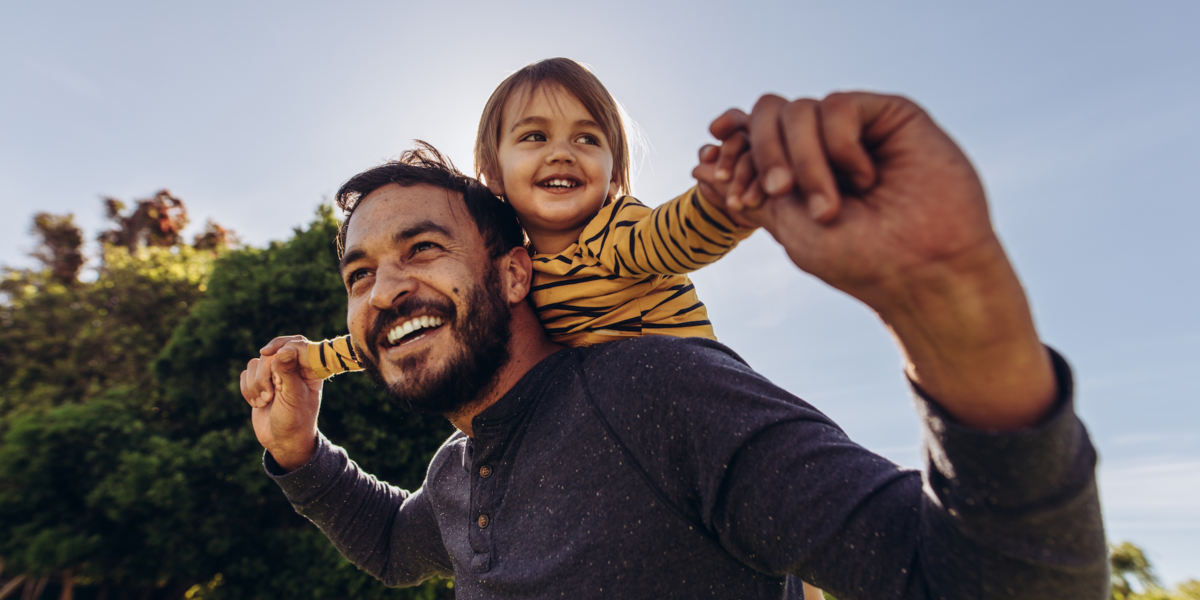 A parent smiles while walking outside with their child on their back