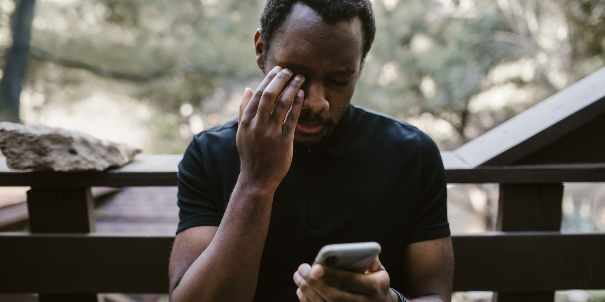A man sits on a deck outside and looks at his phone while holding his head, as if overwhelmed or disappointed