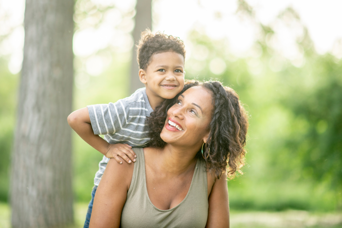 A son climbs on his mother's back in front of tree trunks and greenery. They smile and wear a T-shirt and a tank top.