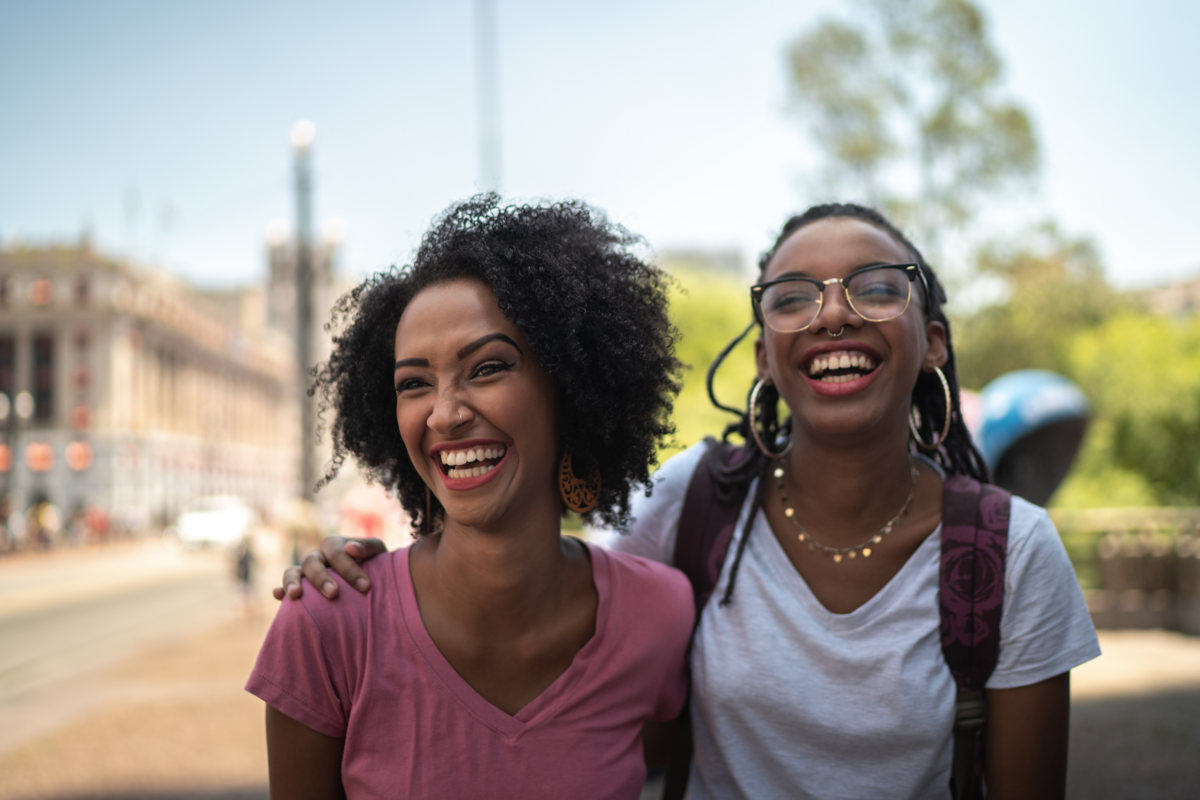 A mother's teenage child, who wears a backpack, puts their arm around her as they both smile and laugh