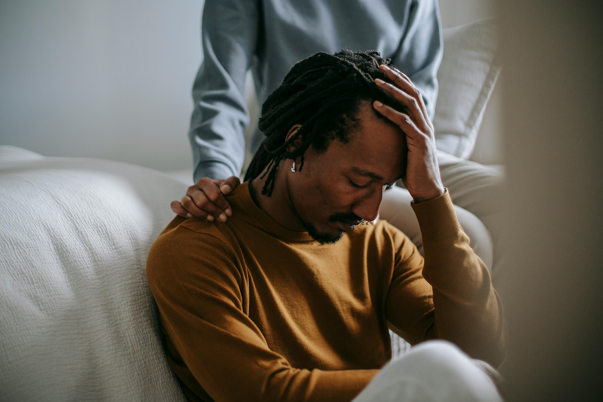 A Black adult with dreads, earrings, and a septum piercing sits in on a couch in sorrow with his head in his hands, while another person rests their hand on their shoulder.