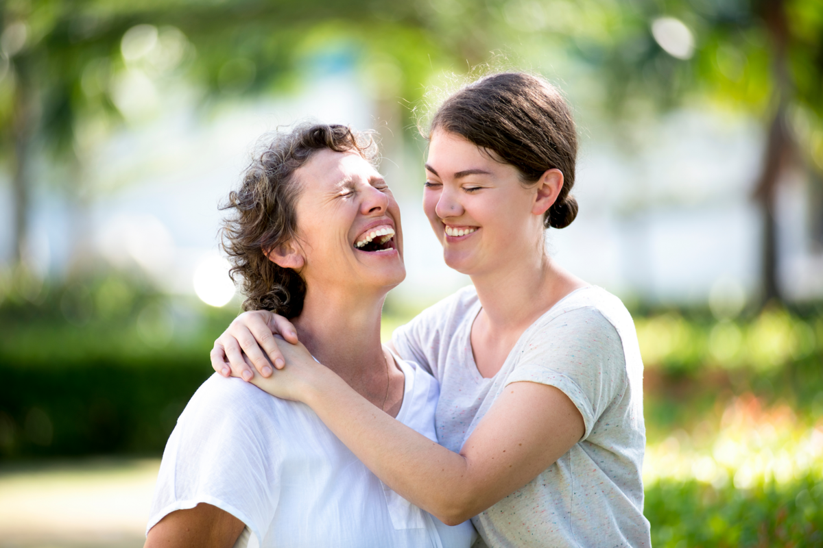 A mother and teenage daughter wear T-shirts and laugh together under the shade of trees.