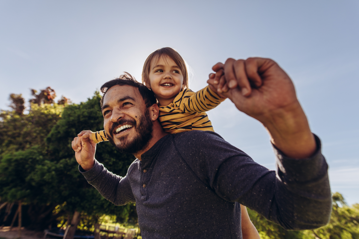 A parent smiles while walking outside with their child on their back
