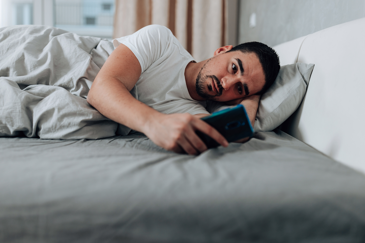 A person lies on their side in bed with the lights on, staring at their phone