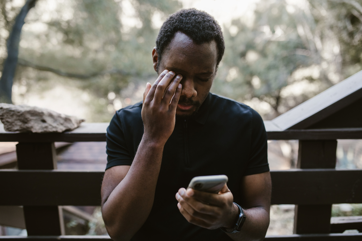 A man sits on a deck outside and looks at his phone while holding his head, as if overwhelmed or disappointed