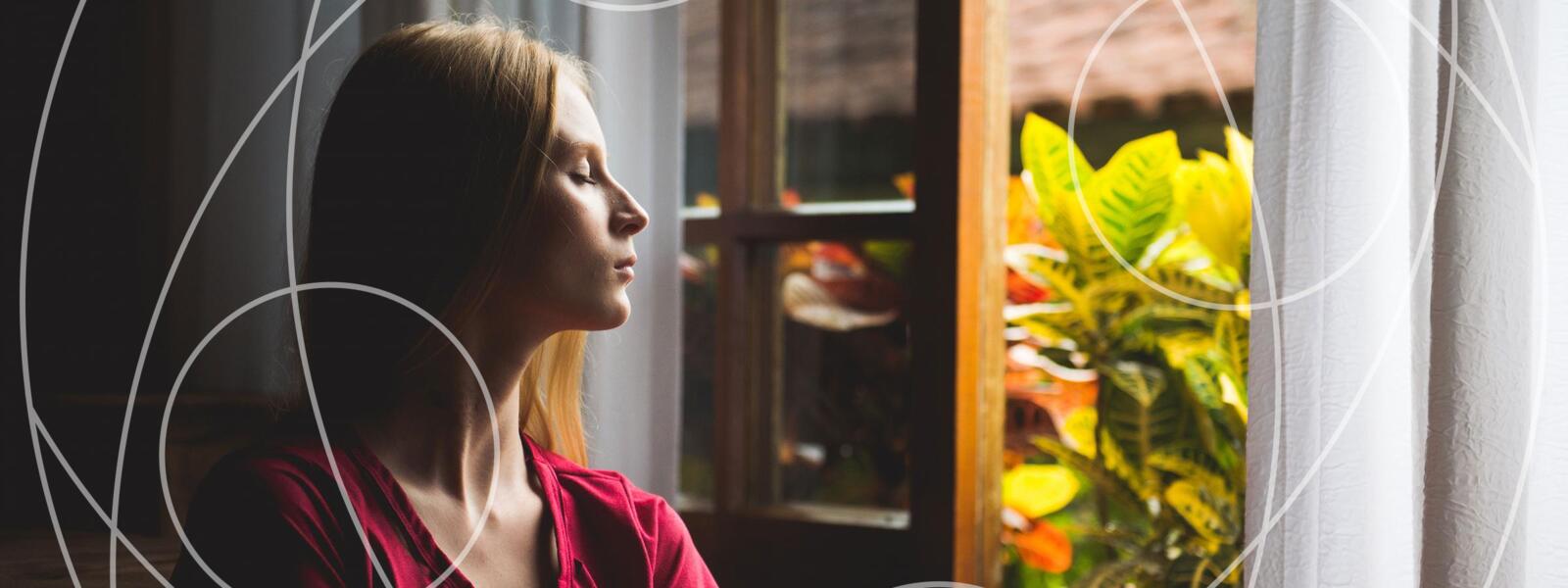 Woman sits by an open window, eyes closed, meditating