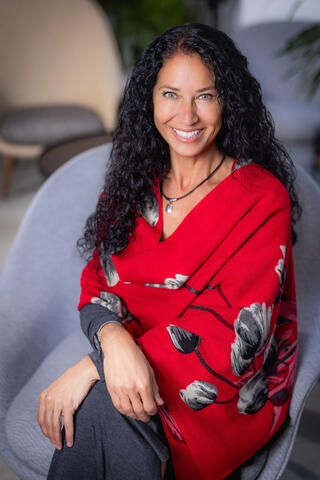 Reem Cutinello wears a red top and sits in a chair, smiling.
