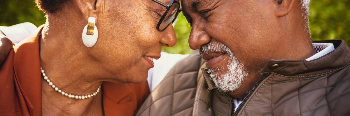 An elderly African American couple smiles and sits closely with their foreheads touching.