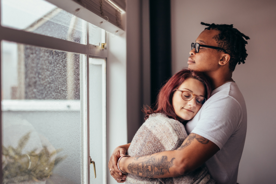 A biracial couple hugs next to a window
