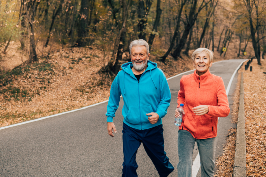 An older couple jogs next to each other along a forest-lined road 