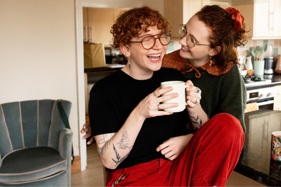 A queer couple laughs together while one puts their arms around the other