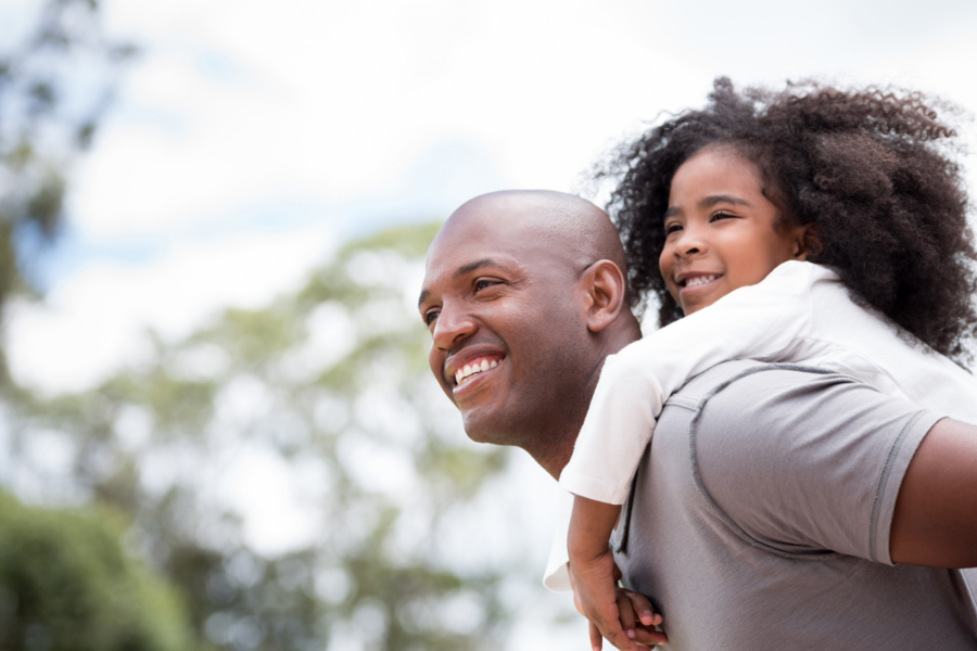 An African-American father smiles while giving his child a piggy back ride