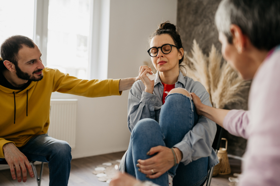 A person sits in a support group setting and closes their eyes while holding a tissue. Others reach their hands out gently.