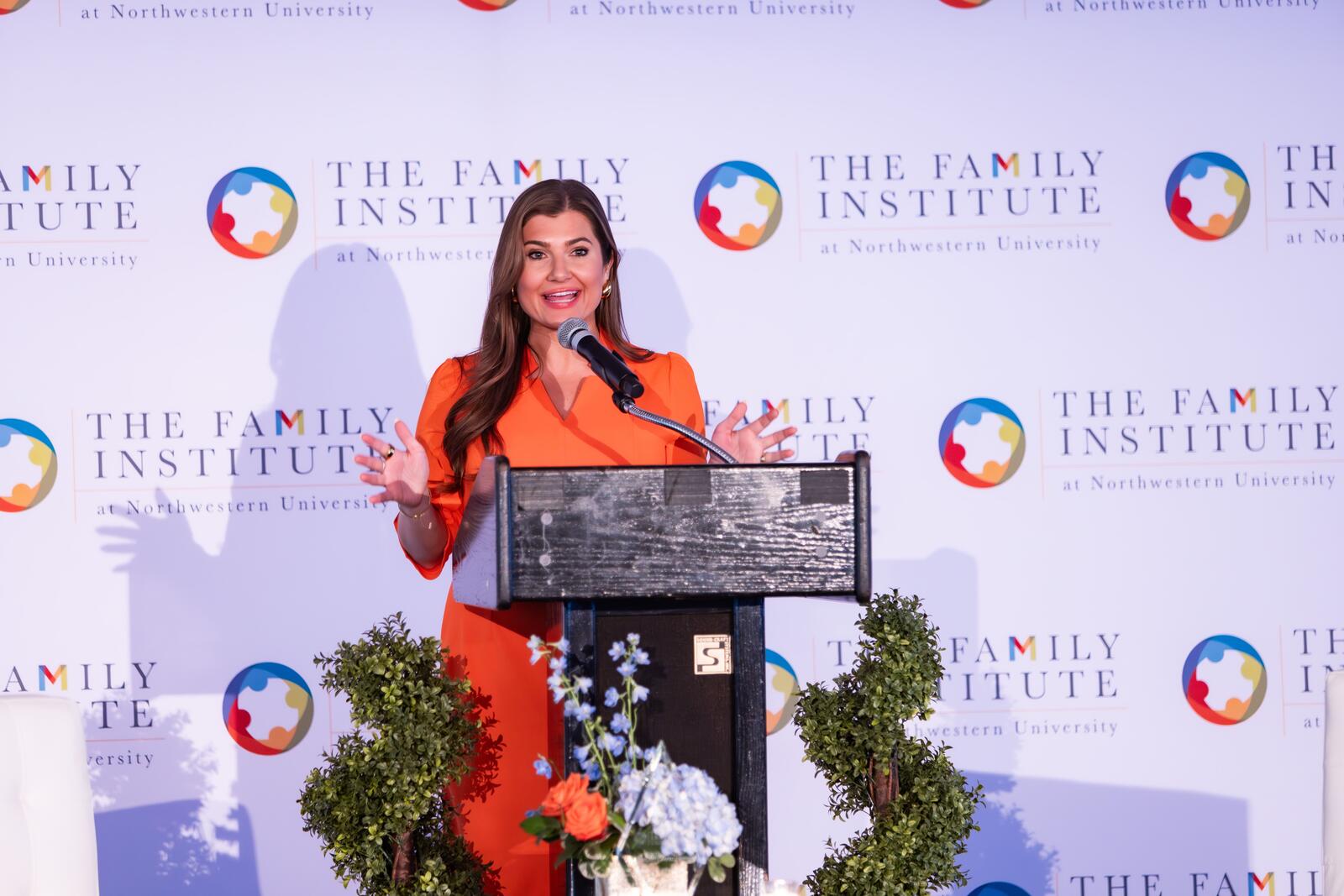 Lexi Sutton from NBC 5 Chicago wears an orange dress and speaks into a microphone at a podium on stage with The Family Institute's logo in the background.