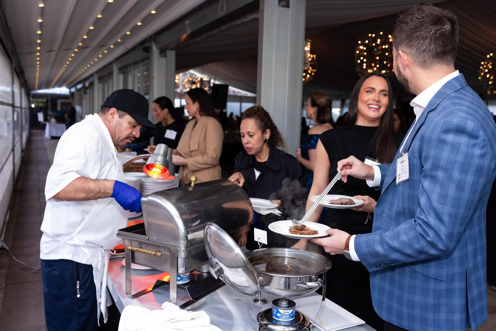 Guests of the Gratitude Gala stand in line at a food station and fill their plates with prime rib and pasta.