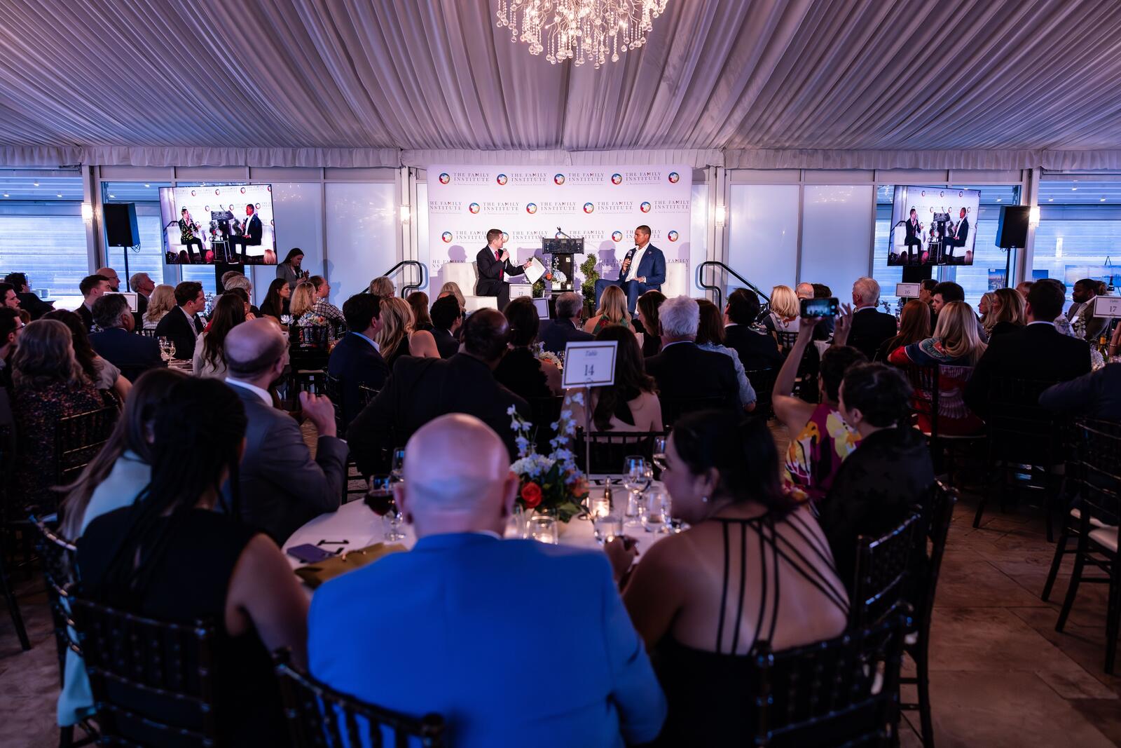 In a crowded room with tables filled with guests, Dr. Jonathan Sutton and Ryan Poles sit on the lit stage and have a discussion.