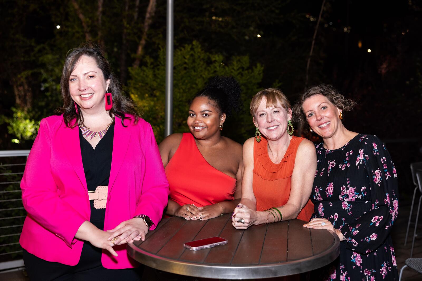 Four guests wearing shades of pink and red smile while standing at a table on the patio at the Gratitude Gala.