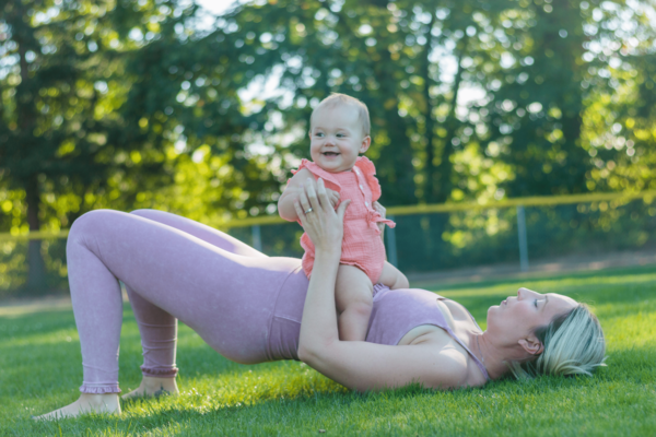 A parent lies in the grass wearing athletic clothing and performing a bridge pose while holding their baby up on their stomach.