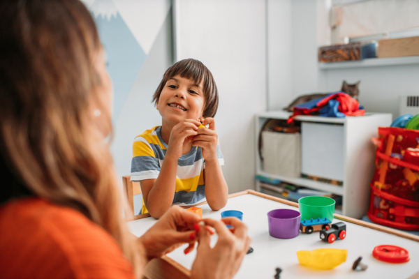 A child sits at a small table in their bedroom or playroom scattered with figurines, cups, and wooden play trains. The child smiles at an adult who also sits at the table.