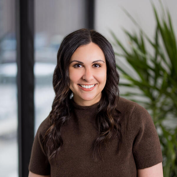 Sophia Odeh smiles and stands next to a chair with her hands placed in front of her on a table and a potted green palm behind her.